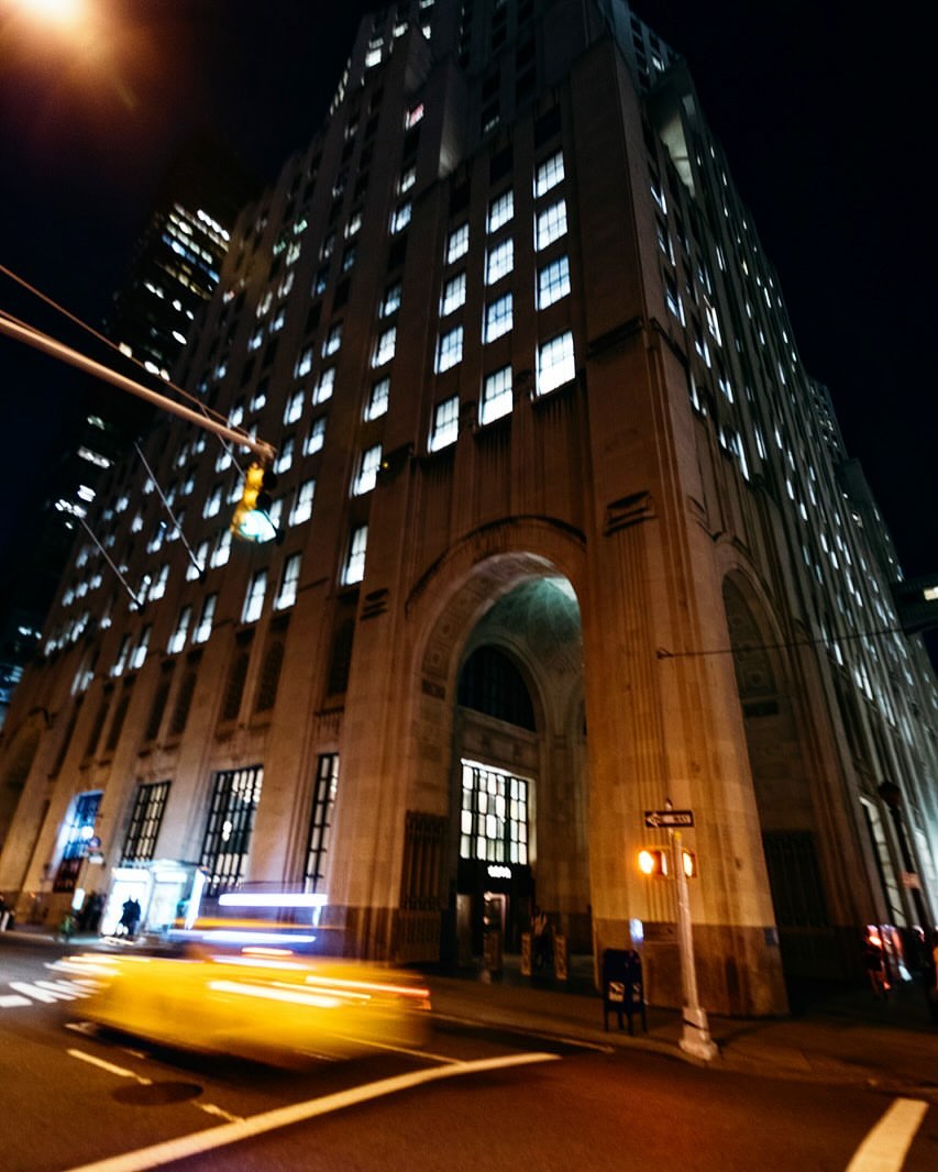 Night scene of a vintage office building with bright windows, an arched entrance, street lights, and blurry taxi cab.