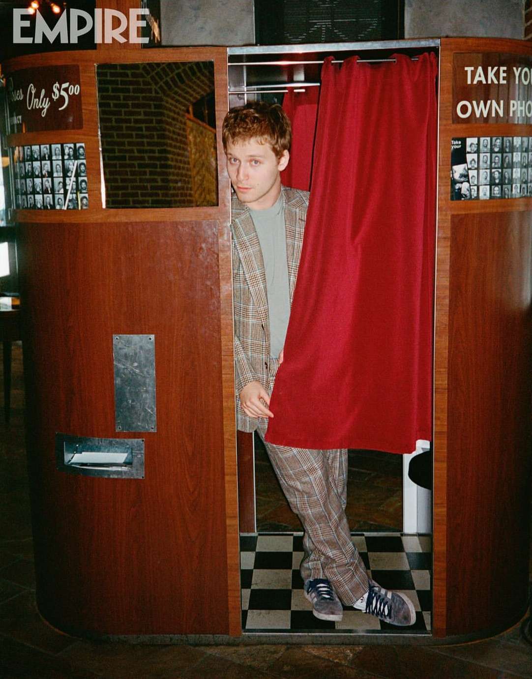 Young man with red hair in a plaid suit posing in a wooden photo booth with red curtains and a black-and-white checkered floor.