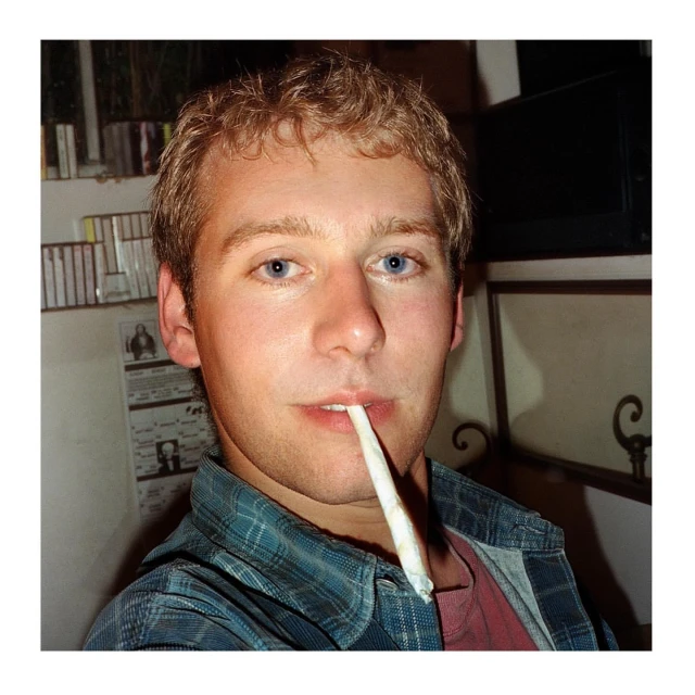 Blond adult male portrait. Indoors, he looks at the camera while a white object sits in his mouth, wearing a blue plaid shirt.