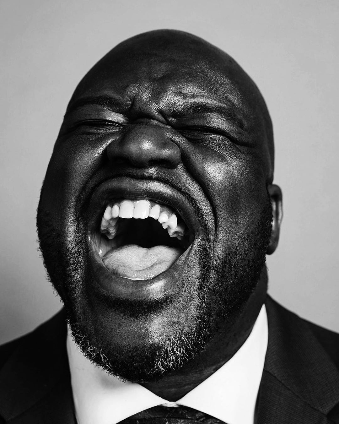 Black and white portrait of an African American man in a suit, eyes closed, mouth open in a shout, against a plain background.