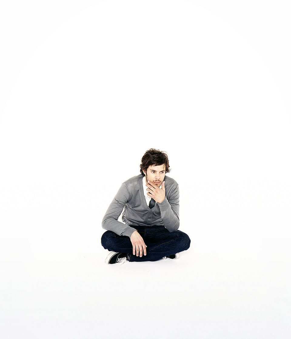 A man in jeans and a grey cardigan sits cross-legged on a white surface, hand to chin, looking off to the side, pensive expression.