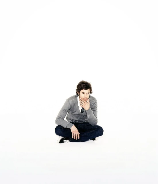 A man in jeans and a grey cardigan sits cross-legged on a white surface, hand to chin, looking off to the side, pensive expression.