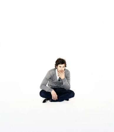 A man in jeans and a grey cardigan sits cross-legged on a white surface, hand to chin, looking off to the side, pensive expression.