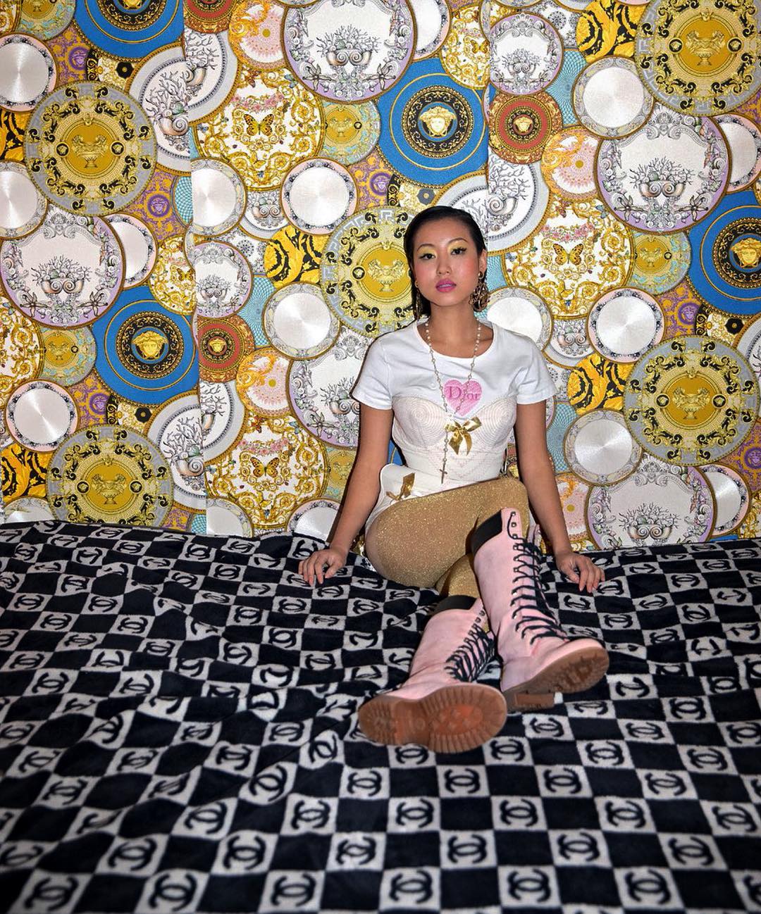 Asian woman sitting on logo-patterned fabric with a designer-patterned wallpaper backdrop, wearing light colored boots and matching top.