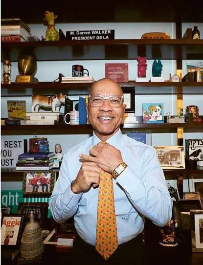 Smiling African-American man in glasses and tie stands in front of bookshelves with books and decorative objects in an indoor setting.
