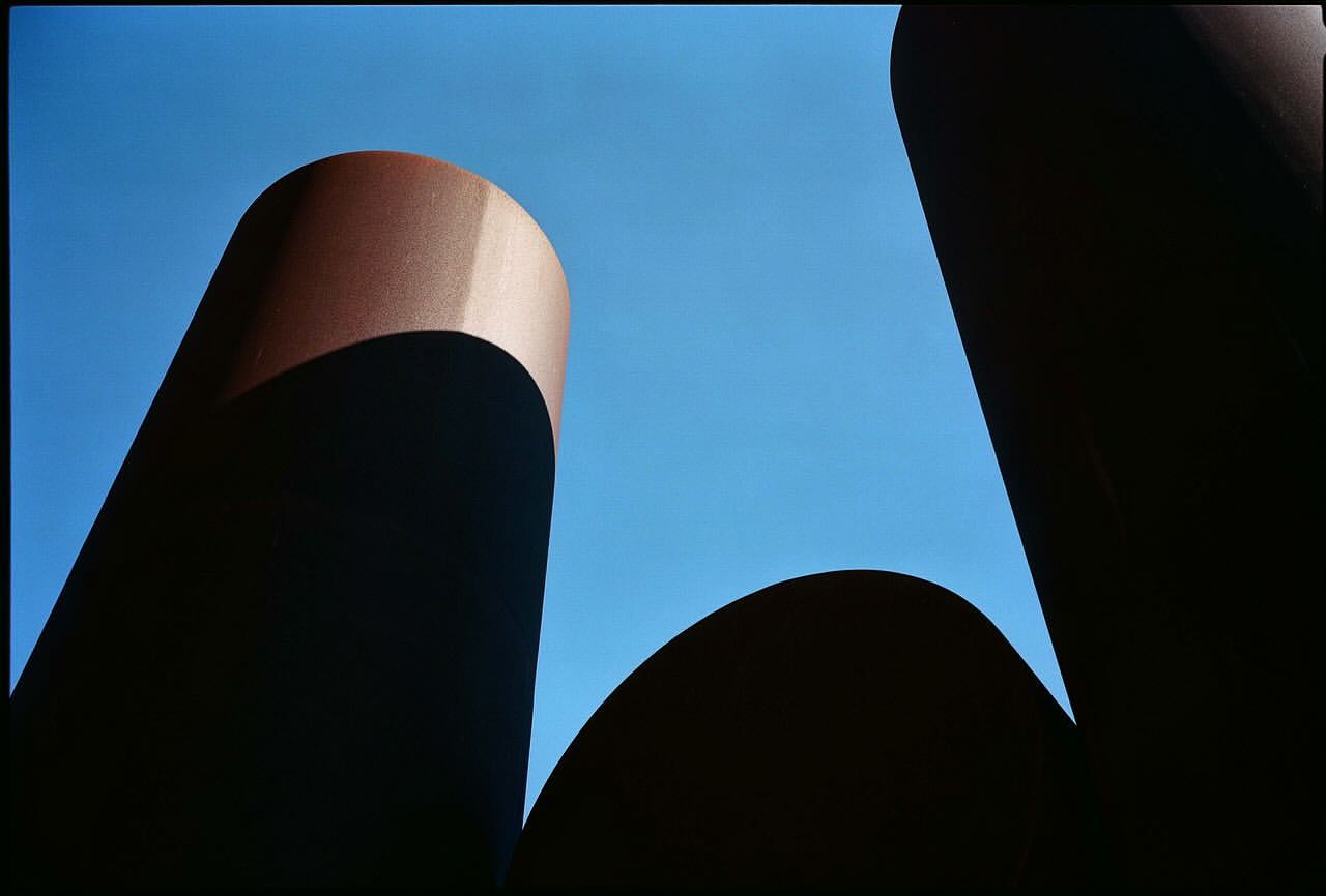 Abstract outdoor shot of dark brown cylindrical structures against a bright blue sky, creating a stark contrast and geometric composition.