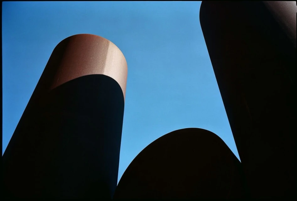 Abstract outdoor shot of dark brown cylindrical structures against a bright blue sky, creating a stark contrast and geometric composition.