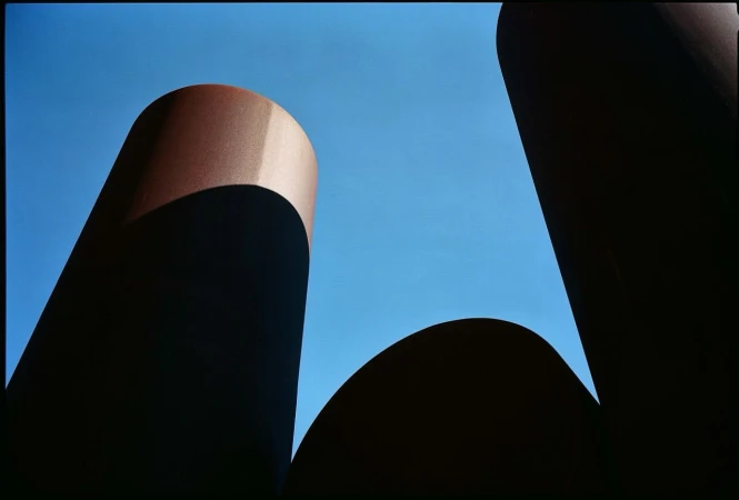 Abstract outdoor shot of dark brown cylindrical structures against a bright blue sky, creating a stark contrast and geometric composition.