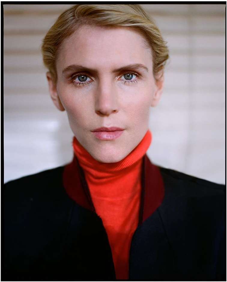 Close-up portrait of a blonde adult woman with blue eyes, wearing a red turtleneck sweater and a black jacket in a neutral indoor setting.