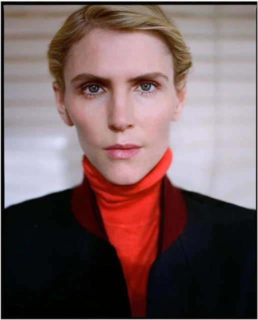 Close-up portrait of a blonde adult woman with blue eyes, wearing a red turtleneck sweater and a black jacket in a neutral indoor setting.