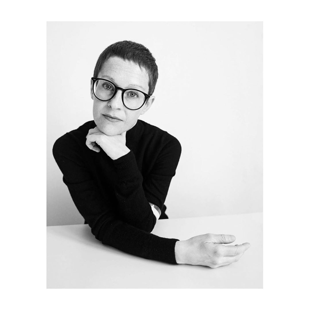 Monochrome portrait of an adult female with short hair and glasses in a black turtleneck, leaning on a white table against a white backdrop.