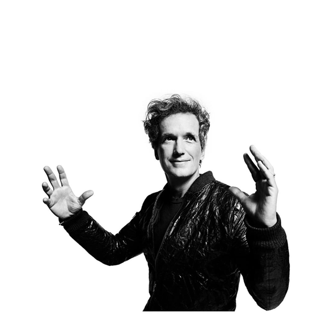 Black and white portrait of a smiling man with curly hair, wearing a dark jacket, with his hands raised against a plain white background.