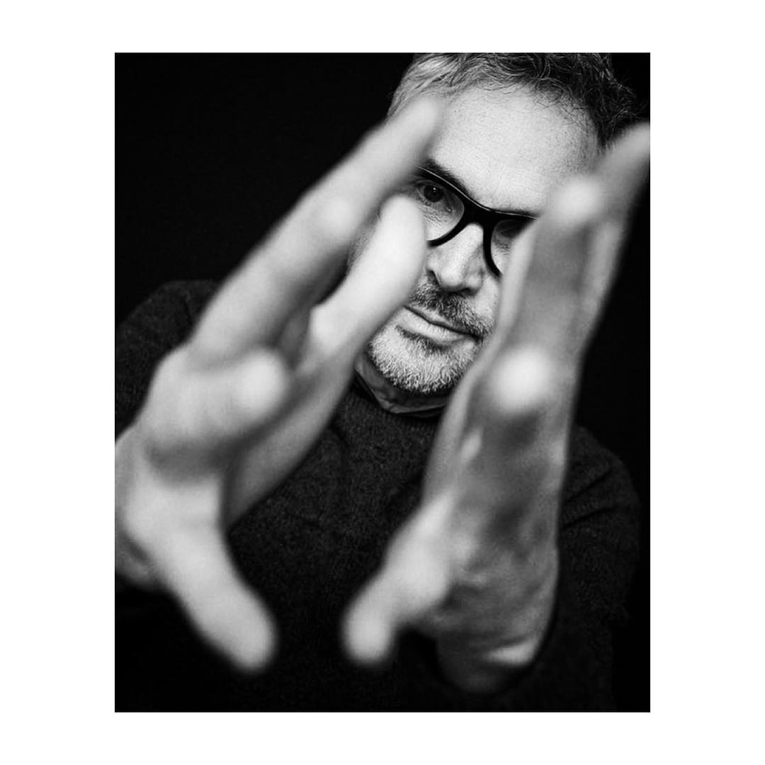 Monochrome portrait of a mature man with glasses, hands raised in foreground, capturing a dramatic, close-up view with dark background.