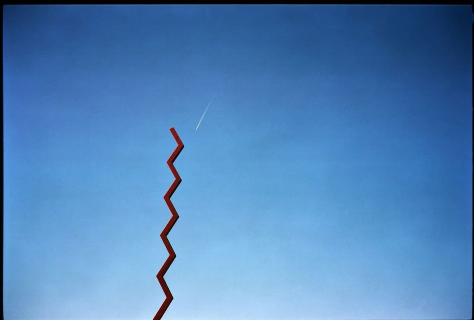 Minimalist outdoor art installation featuring a tall, red zigzag sculpture against a clear blue sky with an airplane contrail above.
