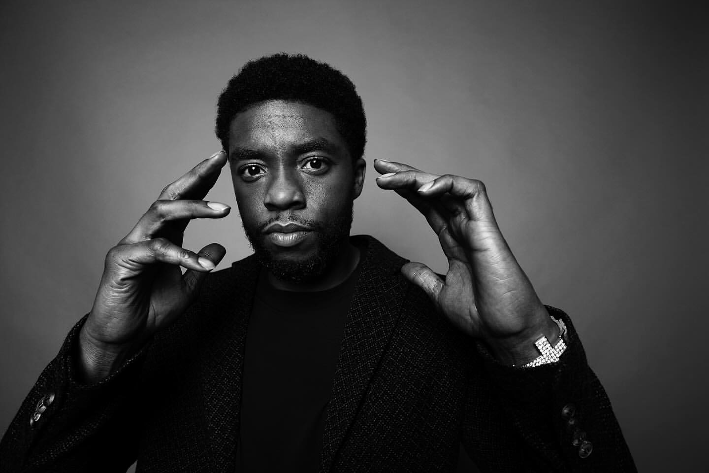 Black and white portrait of Chadwick Boseman in a dark jacket, hands near his temples, against a dark gray background.