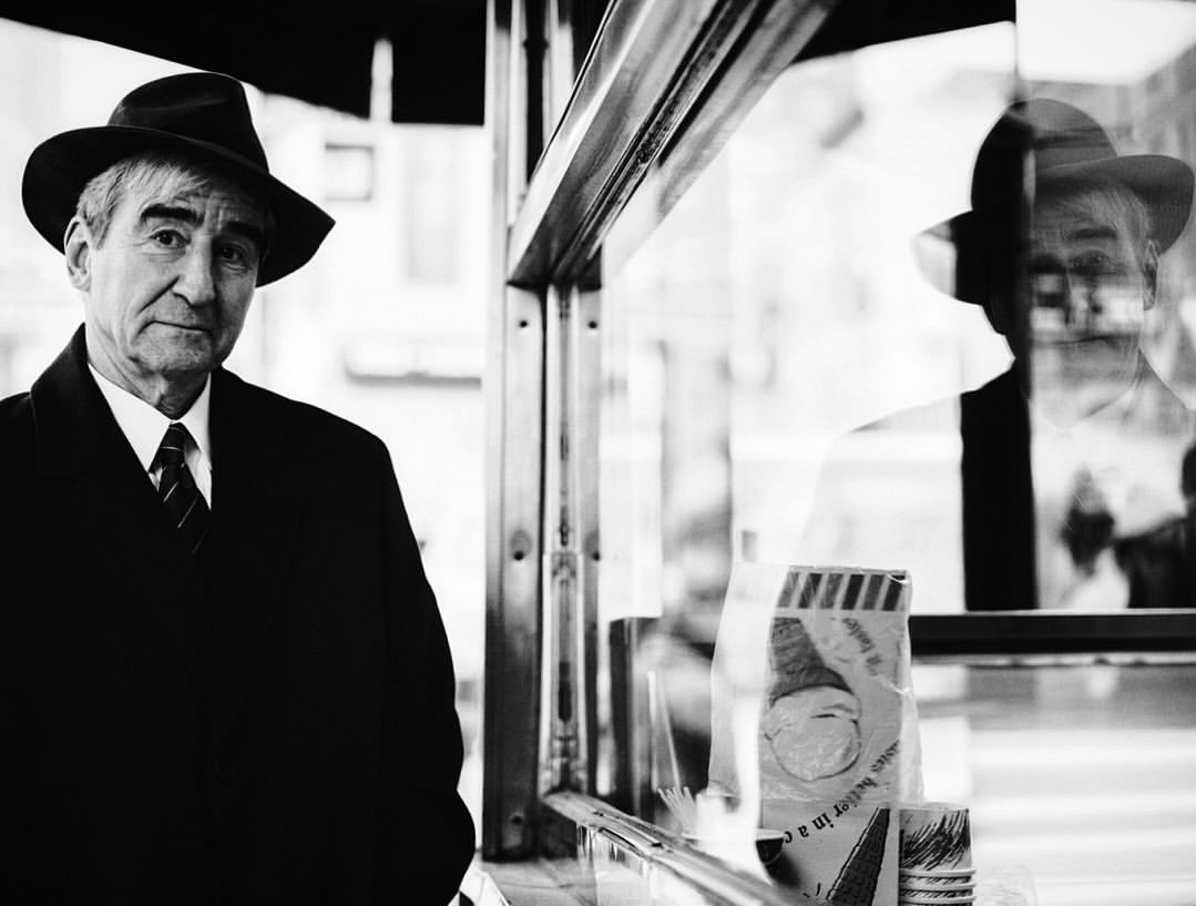 Black and white portrait of an elderly man wearing a fedora and coat, reflected in a glass window with advertisements.