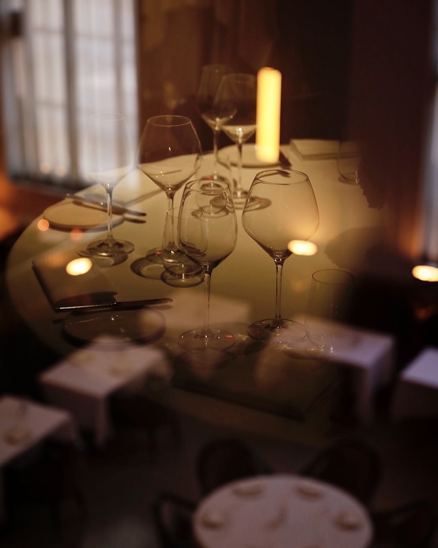 An indoor restaurant scene with a long exposure effect showcases set tables with empty wine glasses and soft lighting from pillar candles.