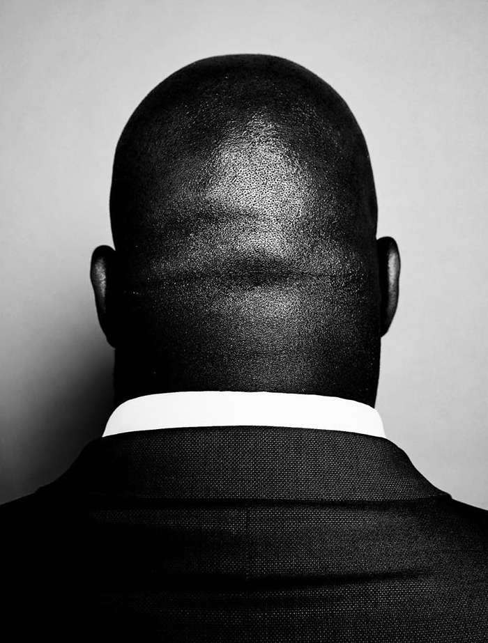 Monochromatic portrait of an African American adult male from behind, showcasing a bald head, dark suit, and white collared shirt.