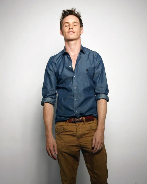 Portrait of a Caucasian adult male with closed eyes wearing a blue collared shirt and khaki pants against a white background in a studio setting.