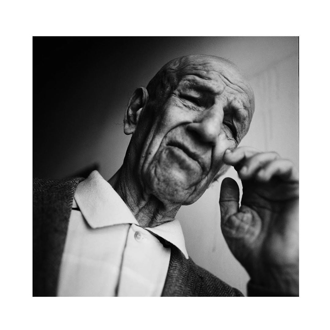 Black and white portrait of an elderly man with wrinkled skin, wearing a collared shirt and cardigan, posing indoors with soft lighting.