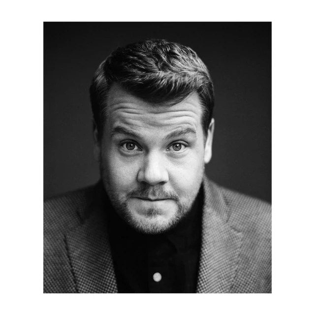 Black and white headshot of adult male with short brown hair wearing a tweed jacket and black shirt against a dark gray background.
