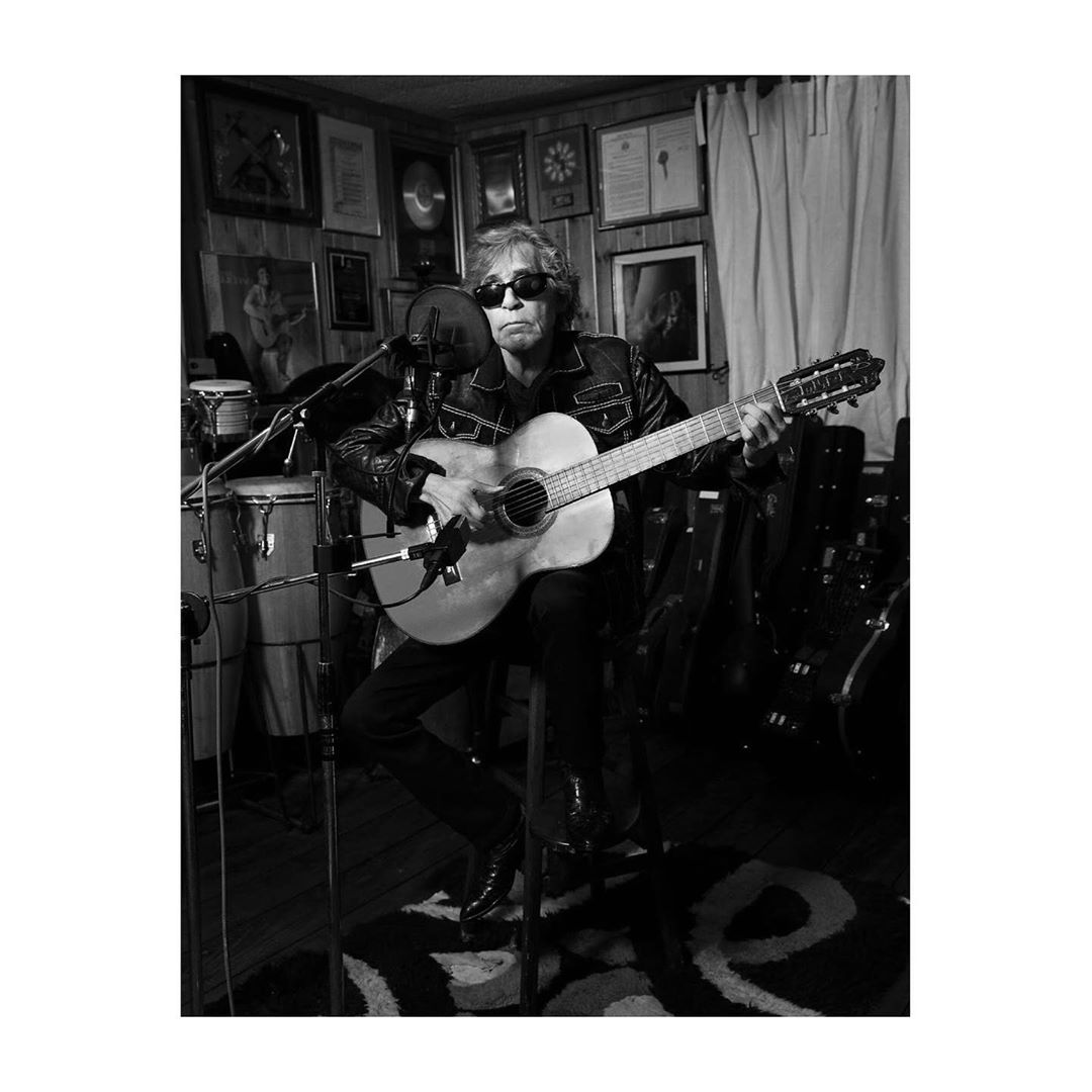 Monochrome shot of a musician playing a twelve-string guitar while seated, wearing a leather jacket, in a wood-paneled room setting.