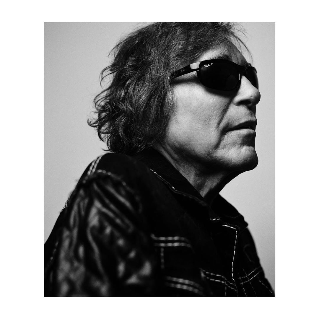 Black and white portrait of an adult man with wavy hair and sunglasses, wearing a leather jacket, against a neutral background.