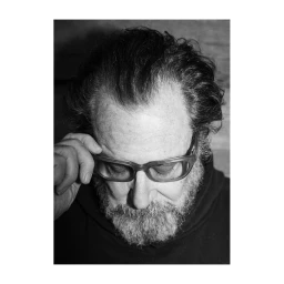 Close-up portrait of a mature man with a beard wearing thick-rimmed glasses and adjusting them, photographed by Jake Chessum.