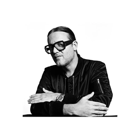 Portrait of a man wearing thick black framed glasses and a dark bomber jacket with arms crossed.