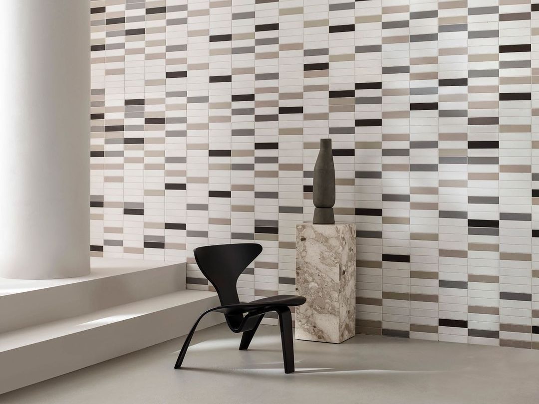 Interior design features modern black chair, sculptural vase, and a wall with horizontally aligned rectangular tiles of various shades.