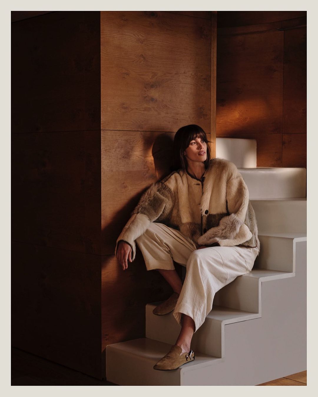 An adult woman with bangs sits on white stairs in front of a wood wall, wearing a light brown and cream jacket and cream pants indoors.