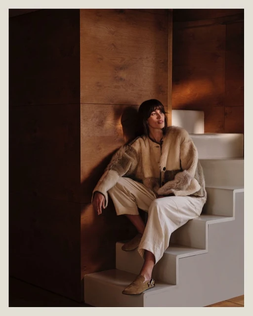 An adult woman with bangs sits on white stairs in front of a wood wall, wearing a light brown and cream jacket and cream pants indoors.