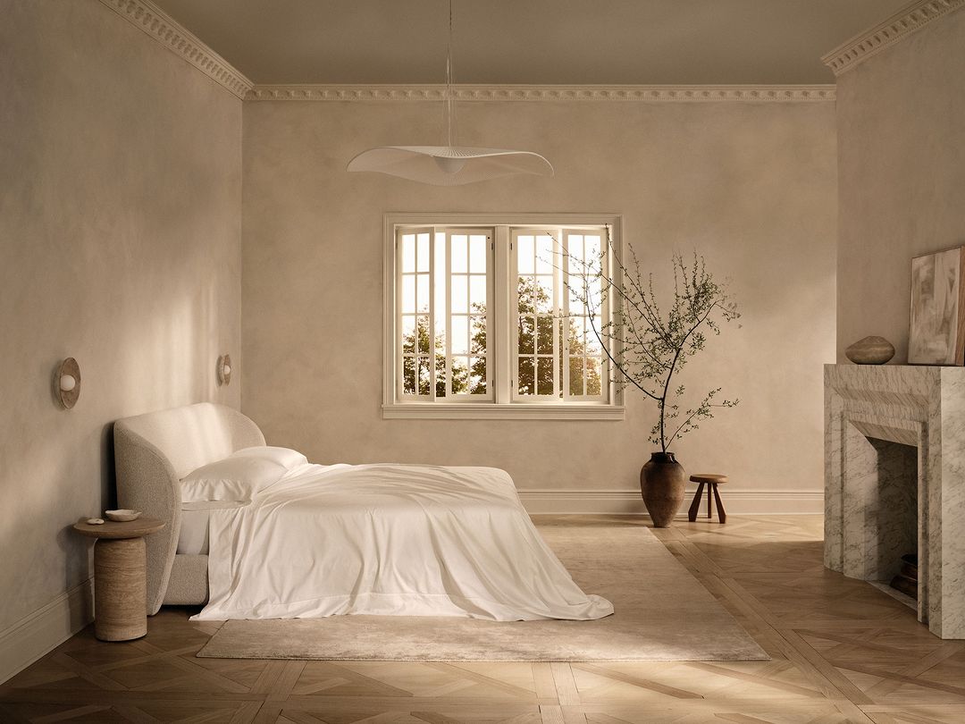 Bright, neutral bedroom with white bedding, marble fireplace, vase with stems, wood floors, and soft light from a large window.