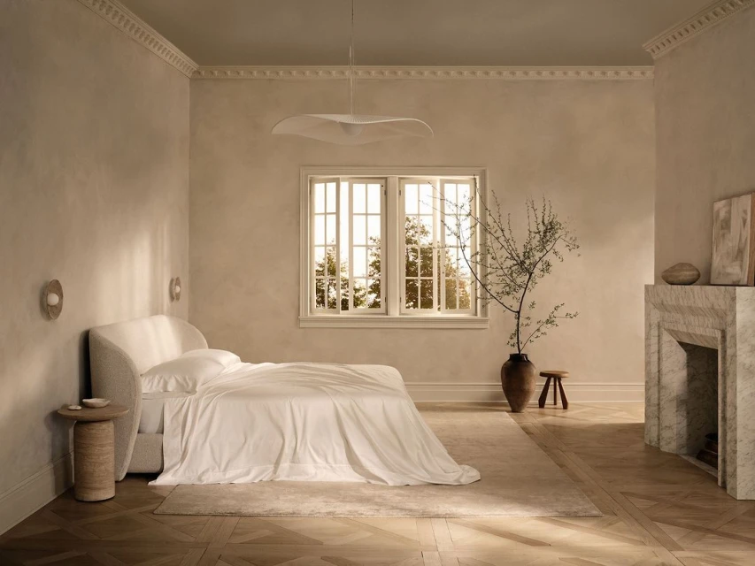 Bright, neutral bedroom with white bedding, marble fireplace, vase with stems, wood floors, and soft light from a large window.