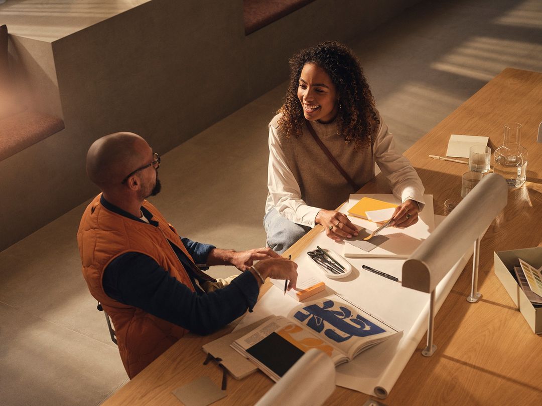 Two people, an adult woman and an adult man, sit at a table with design materials and art books, looking at each other and smiling.