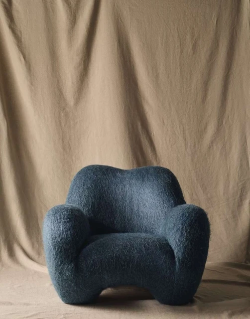Eye-level studio shot of a blue, shaggy armchair against a tan fabric backdrop. Minimalist interior design highlights texture and form.