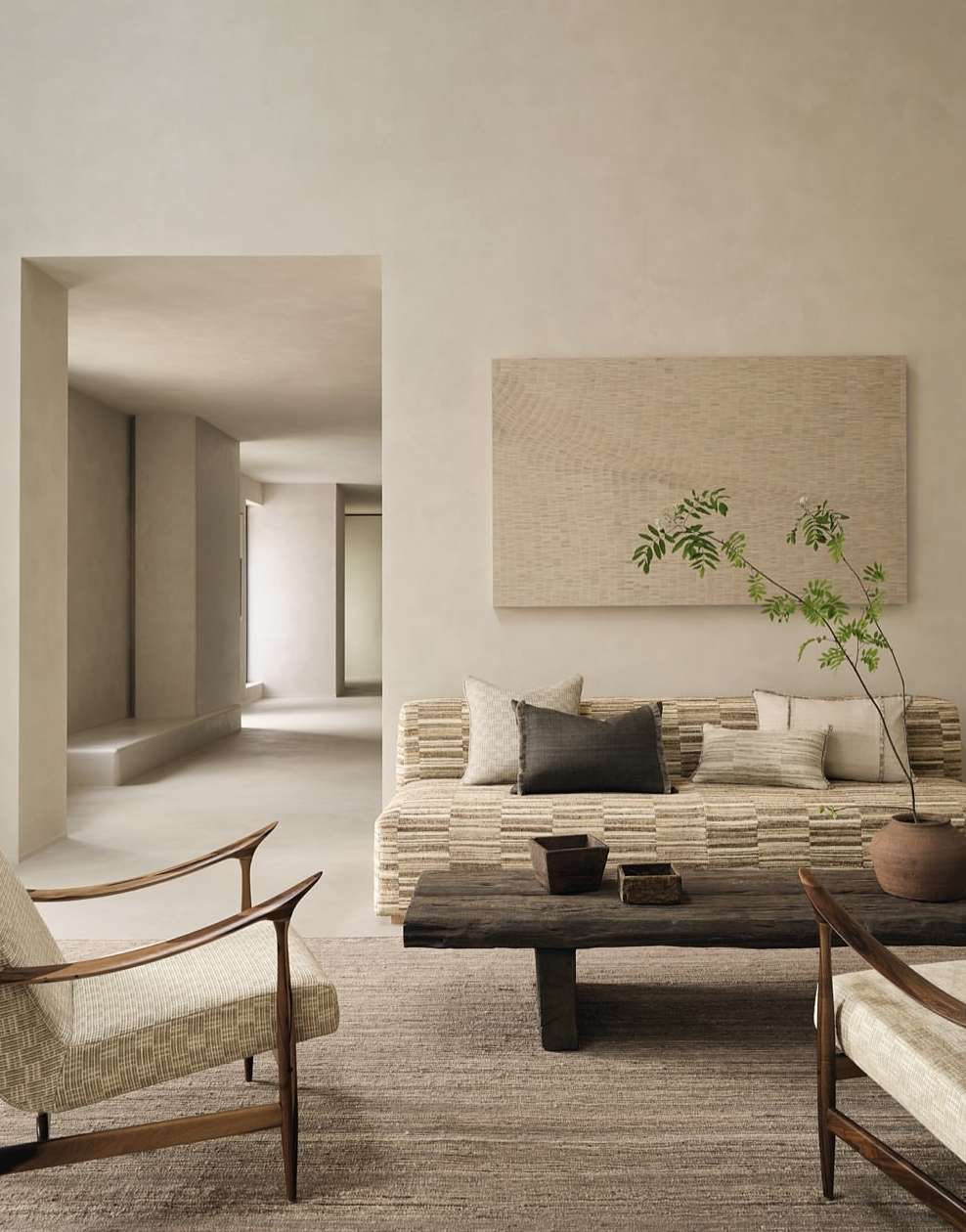 Beige living room with neutral-tone furniture, dark wood coffee table, and artwork above a sofa adorned with cushions and a plant.