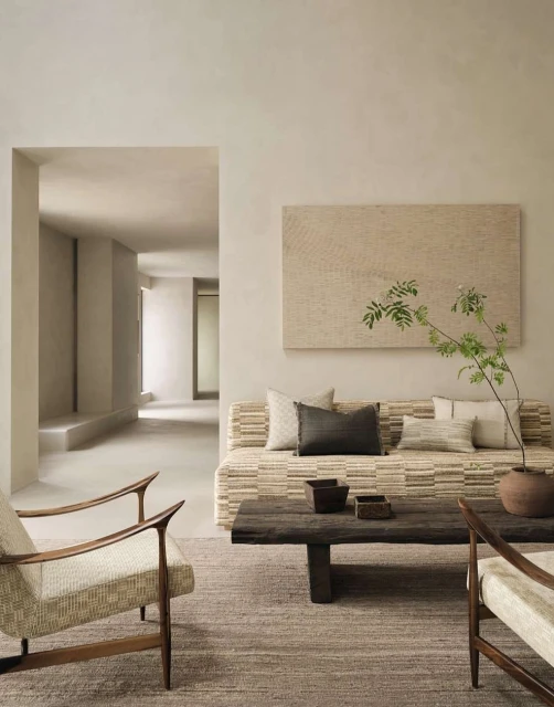 Beige living room with neutral-tone furniture, dark wood coffee table, and artwork above a sofa adorned with cushions and a plant.