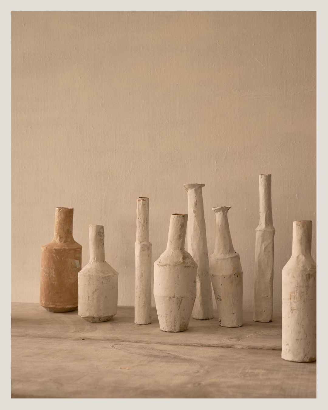 Still life photograph of nine assorted vases in neutral tones, arranged on a table against a light beige textured wall.