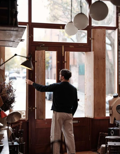 Back view of a man opening wooden shutters in a vintage-style shop, revealing a city street scene, with hanging lamps visible.