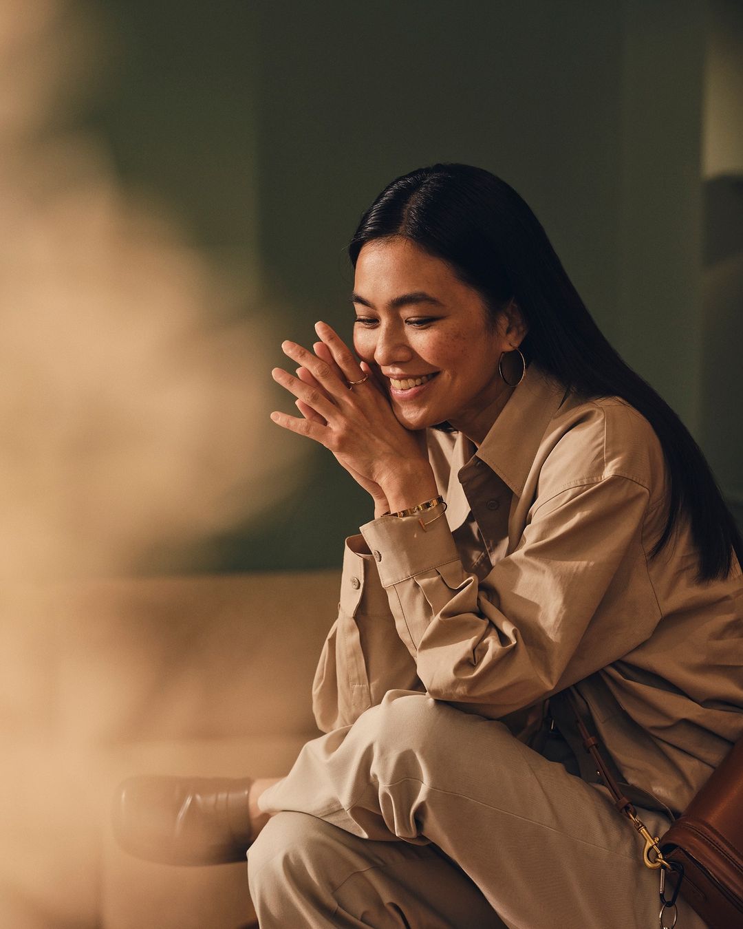 Smiling Asian woman in a beige shirt and pants sits indoors with her hands clasped, showcasing a relaxed pose and elegant hoop earrings.