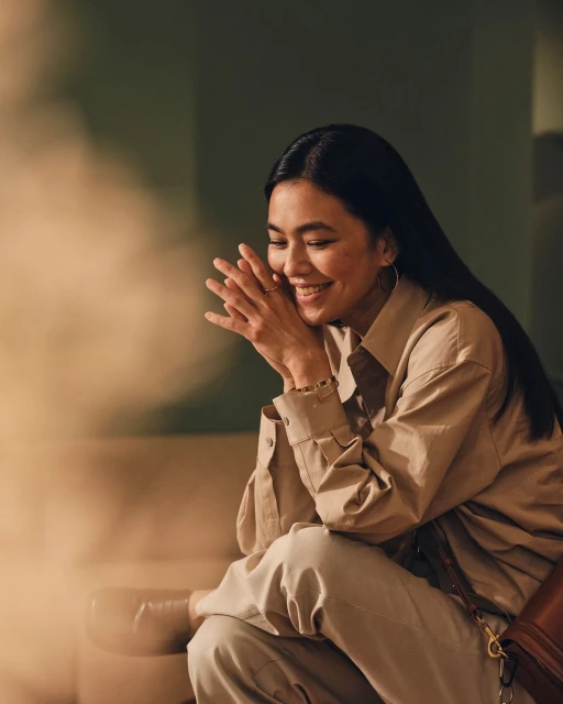 Smiling Asian woman in a beige shirt and pants sits indoors with her hands clasped, showcasing a relaxed pose and elegant hoop earrings.