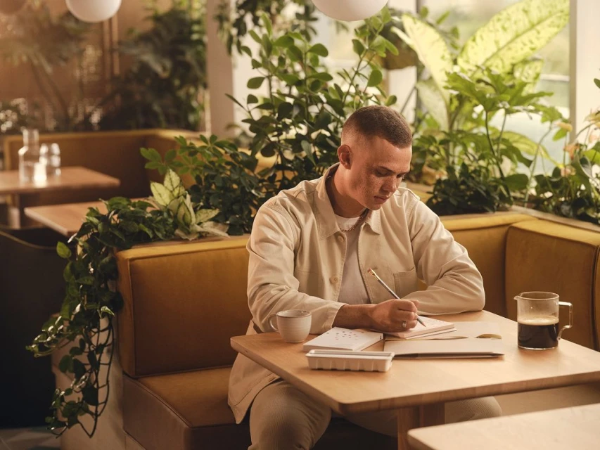 Young adult man sitting at a table in a cafe, writing in a notebook with green plants and bright natural light in the background.
