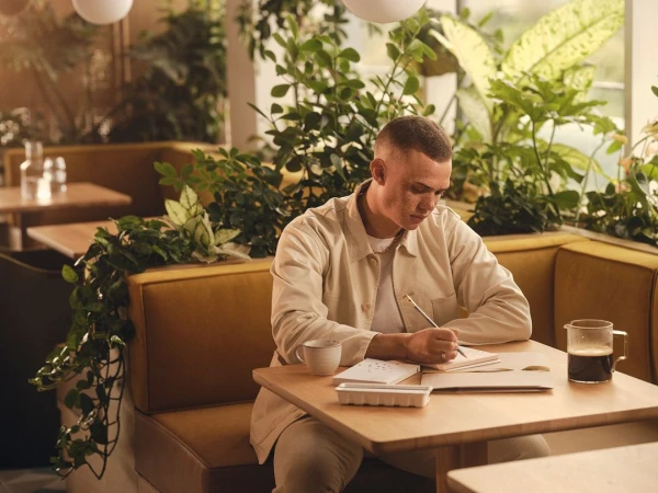 Young adult man sitting at a table in a cafe, writing in a notebook with green plants and bright natural light in the background.
