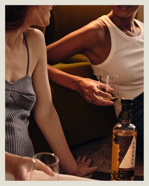 Two women relaxing indoors with an Ardray whisky bottle. One woman holds a glass of whisky. Warm lighting highlights the dark color palette.