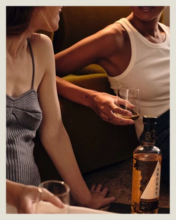 Two women relaxing indoors with an Ardray whisky bottle. One woman holds a glass of whisky. Warm lighting highlights the dark color palette.