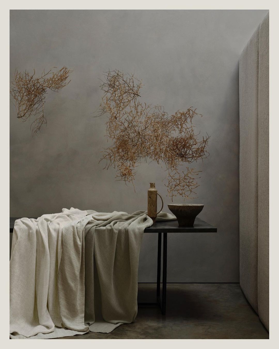 Minimalist interior with a draped linen cloth on a table, a stoneware bowl and bottle, and decorative branches against a neutral wall.