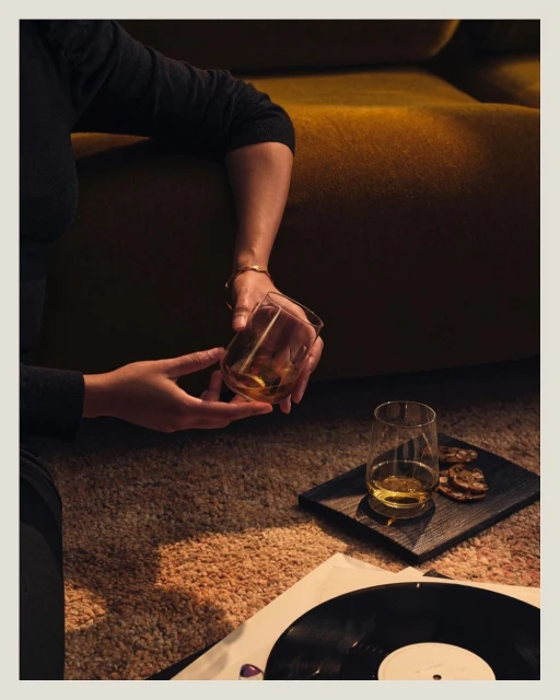 Person holding glass of amber liquid, vinyl record, and snacks on the floor creating a cozy, relaxed indoor atmosphere with warm lighting.