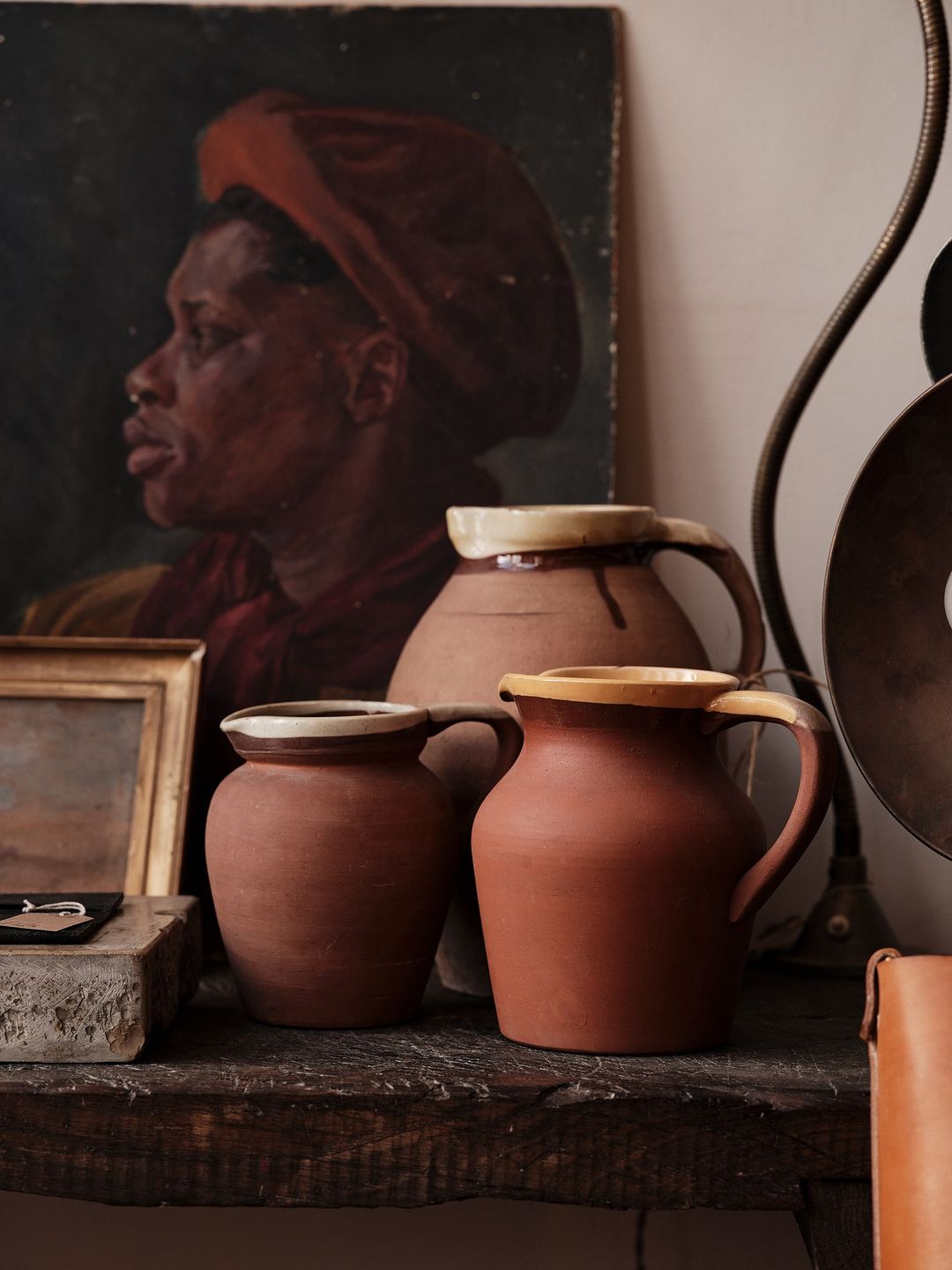 Still life photo features a collection of earthenware pitchers arranged on a dark wooden surface, with a framed portrait in the background.