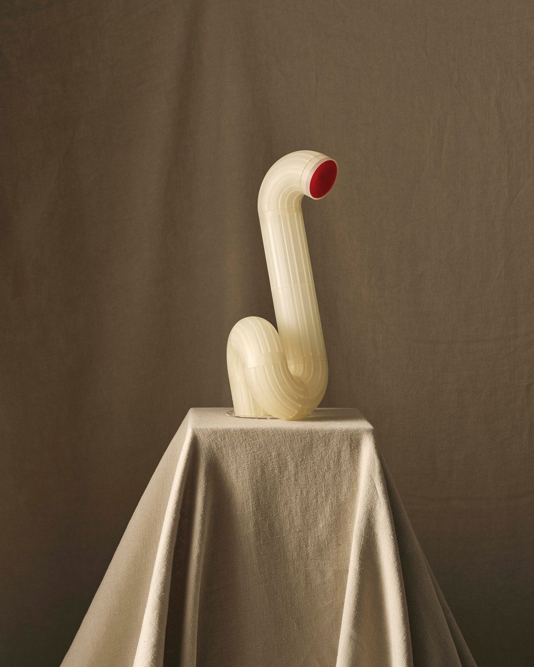 Still life photograph of a contemporary art sculpture on a draped pedestal with an off-white, tan, and red color palette in a photo studio.